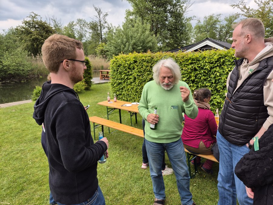 Im Vordergrund unterhalten sich 3 Personen, die auf einer Wiese stehen. Links im Hintergrund fließt ein Fluss, auf der Wiese stehen Biertischgarnituren, auf denen Flaschen sowie Teller  und Besteck stehen und liegen. Im Hintergrund eine grüne Hecke sowie Personen, die an den Biertischen sitzen.