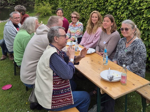 An einer Biertischgarnitur sitzen 10 Personen, auf dem Tisch stehen Getränke und Teller sowie Besteck. Die Biertischgarnitur steht auf einer Wiese, im Hintergrund ist eine grüne Hecke
