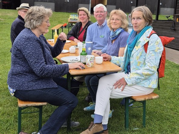 Sechs Personen sitzen an einer Biertisch-Garnitur, die auf einer Wiese steht. Im Hintergrund sieht man ein Holzhaus mit verschiedenen offenen Toren, oben rechts noch eine Girlande mit bunten Lampions